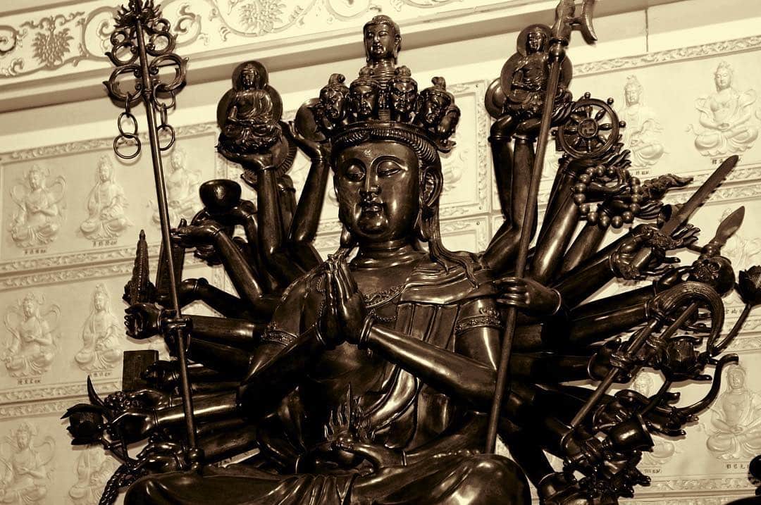 Escultura dorada de Guan Yin de muchos brazos, con un halo de manos abiertas a su espalda, en tonos sepia.
