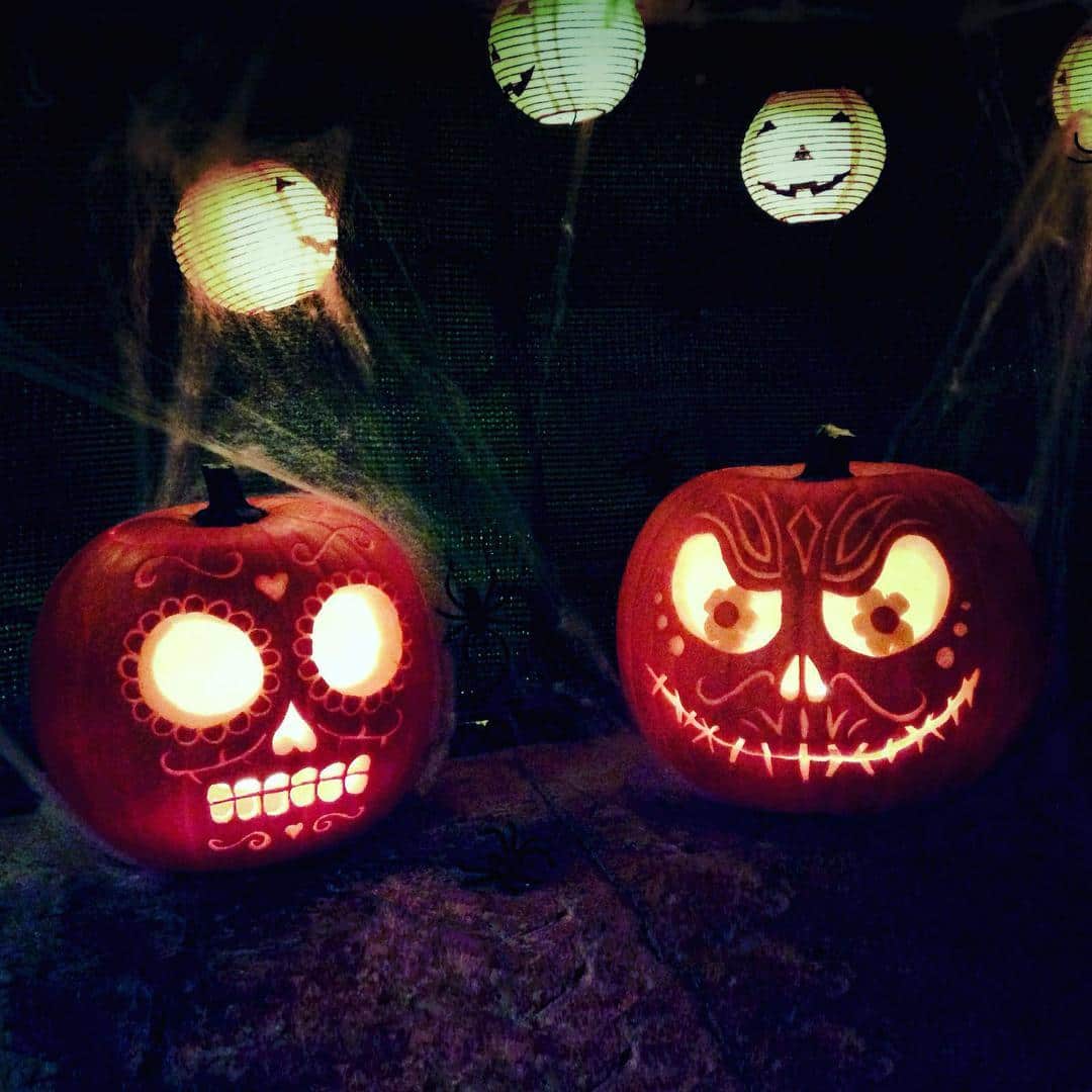 Dos calabazas de Halloween talladas e iluminadas por dentro, junto a farolillos de papel con caras sonrientes, sobre fondo oscuro.