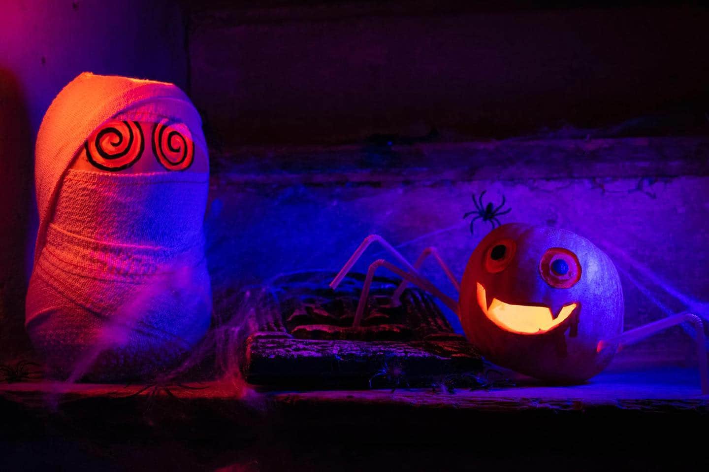 Decoración de Halloween iluminada en morado y naranja, con una calabaza sonriente, una araña y una figura con espirales en los ojos.