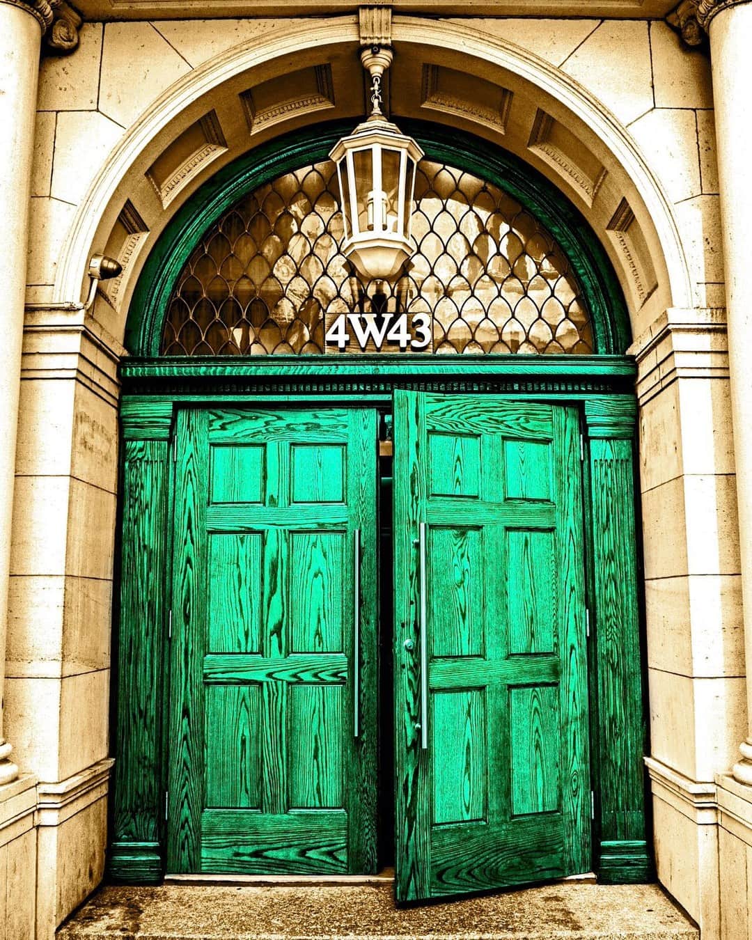 Puerta doble de madera pintada de verde bajo un arco de piedra, con un farol y una reja en el tímpano.