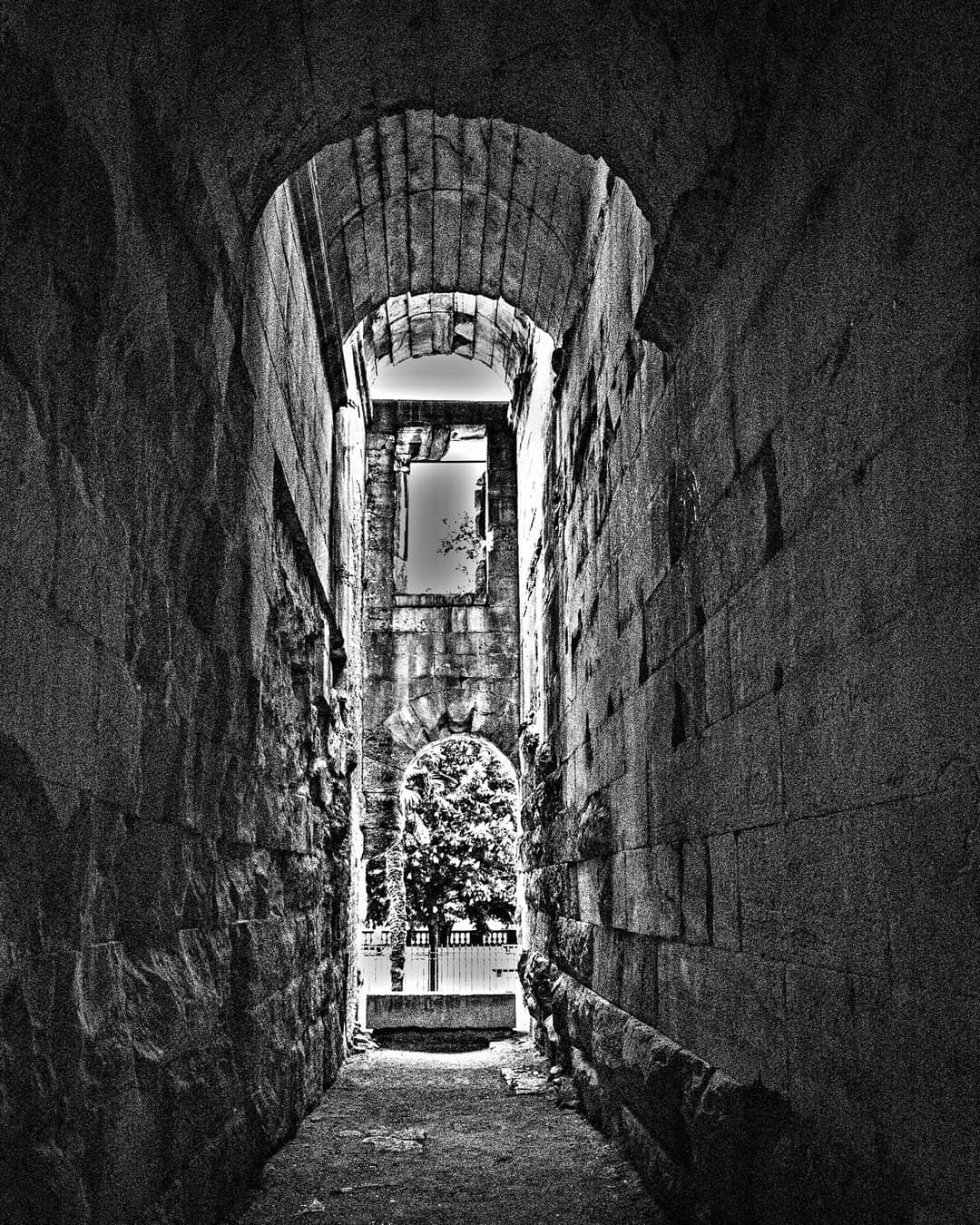 Pasadizo abovedado de piedra que se estrecha hacia una salida iluminada, en blanco y negro.