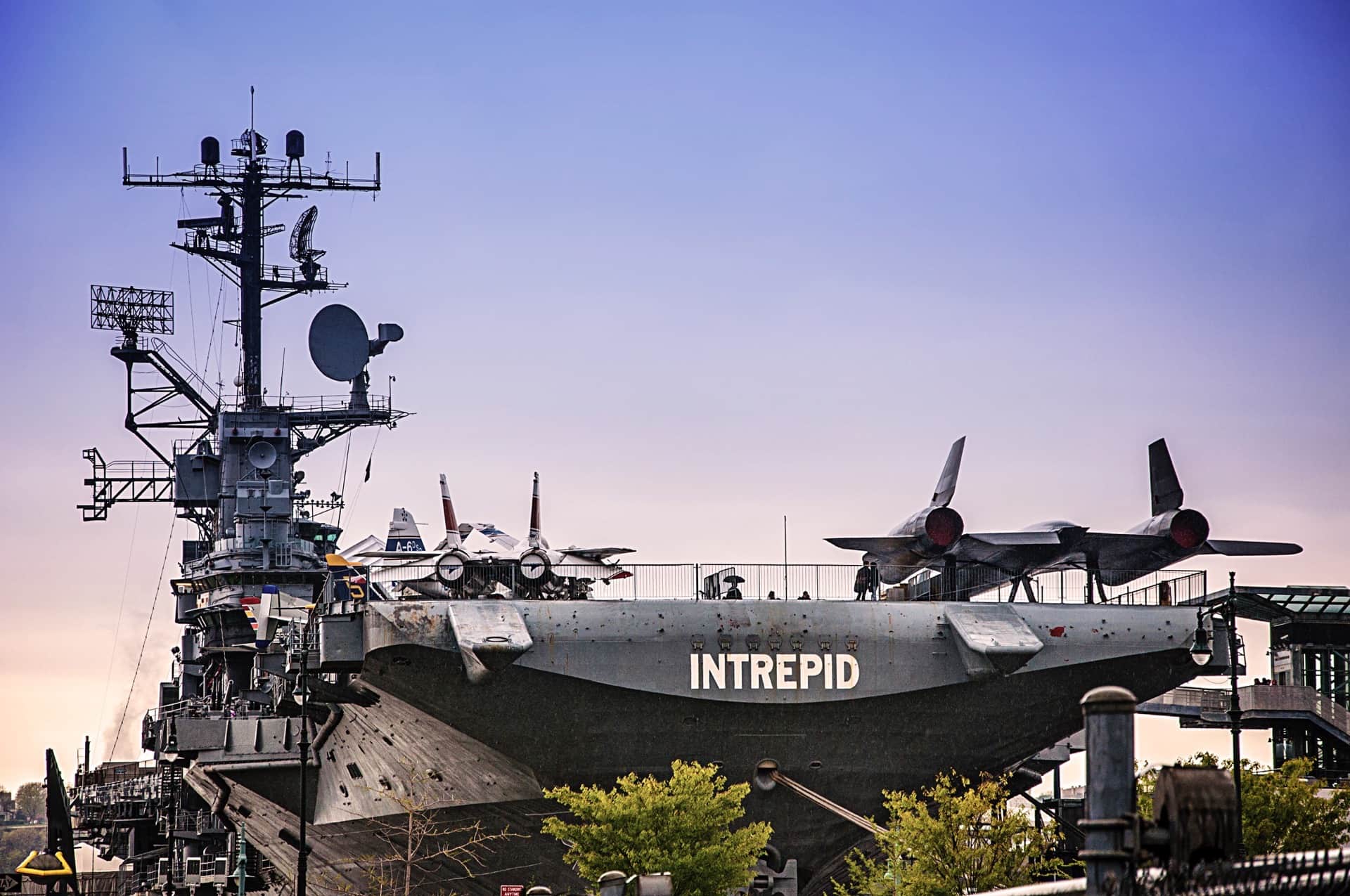 Portaaviones amarrado con el rótulo INTREPID en el casco y aviones en cubierta, bajo un cielo violeta al atardecer