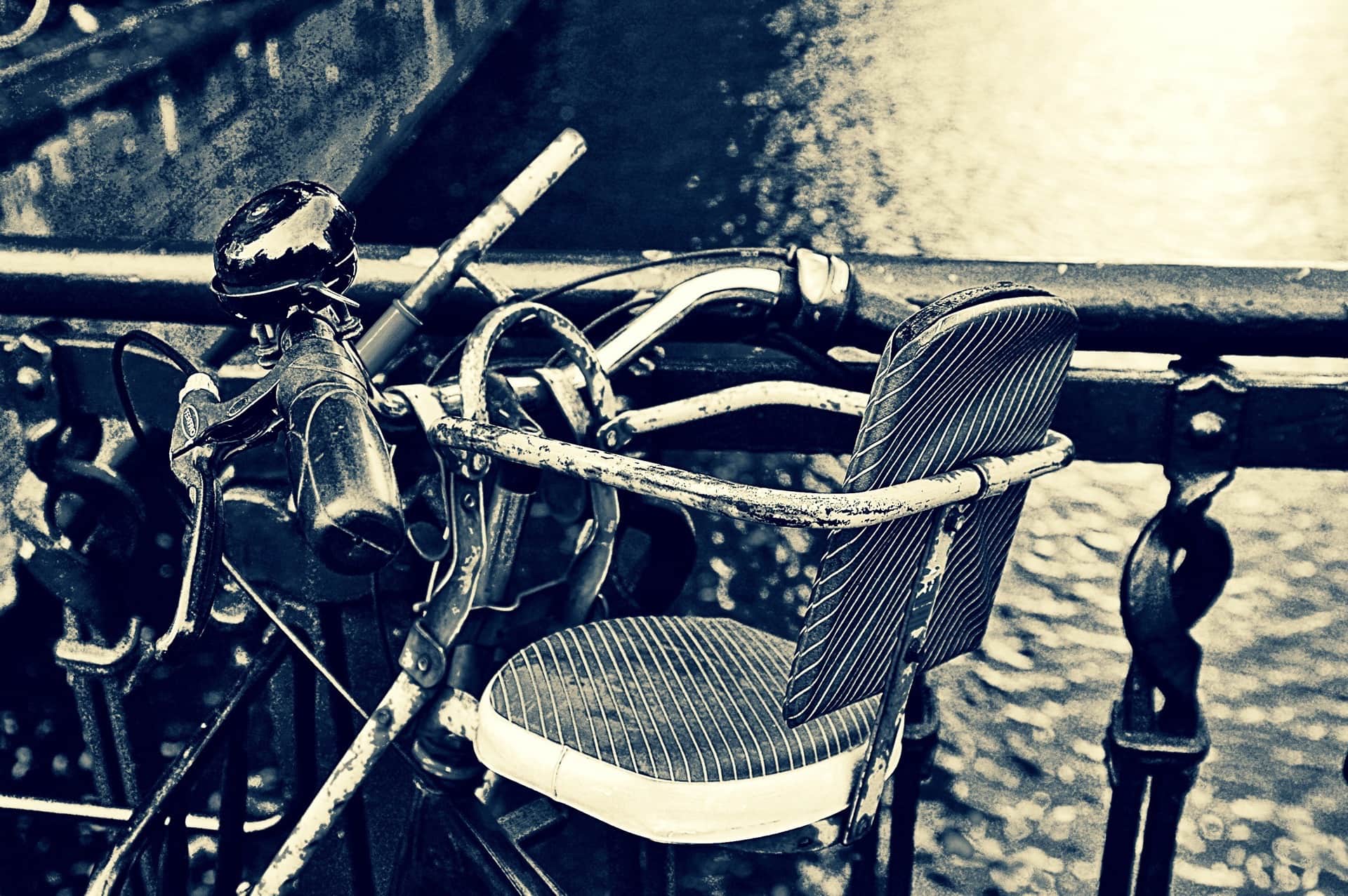 Manillar de bicicleta vieja con timbre y asiento infantil a rayas, apoyada en una barandilla sobre el agua, en tonos azules