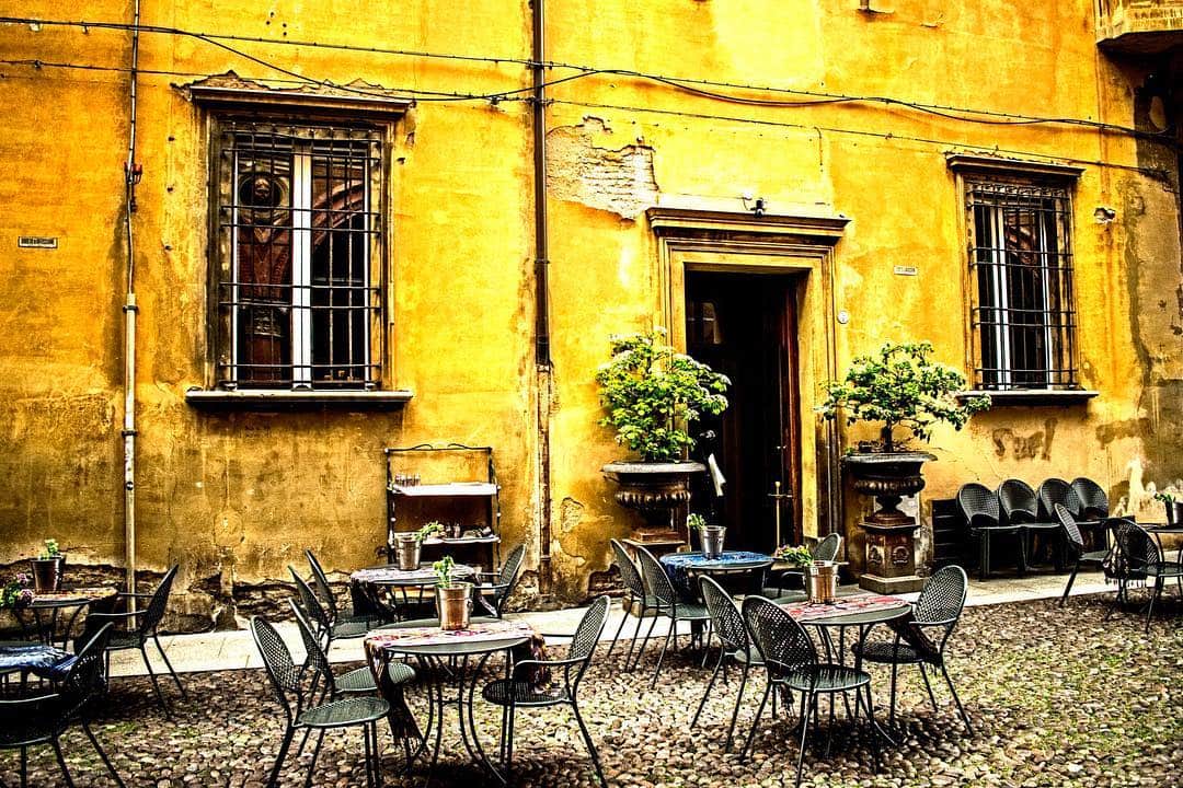 Terraza con mesas y sillas metálicas en un patio, ante una fachada amarilla desconchada con macetas.