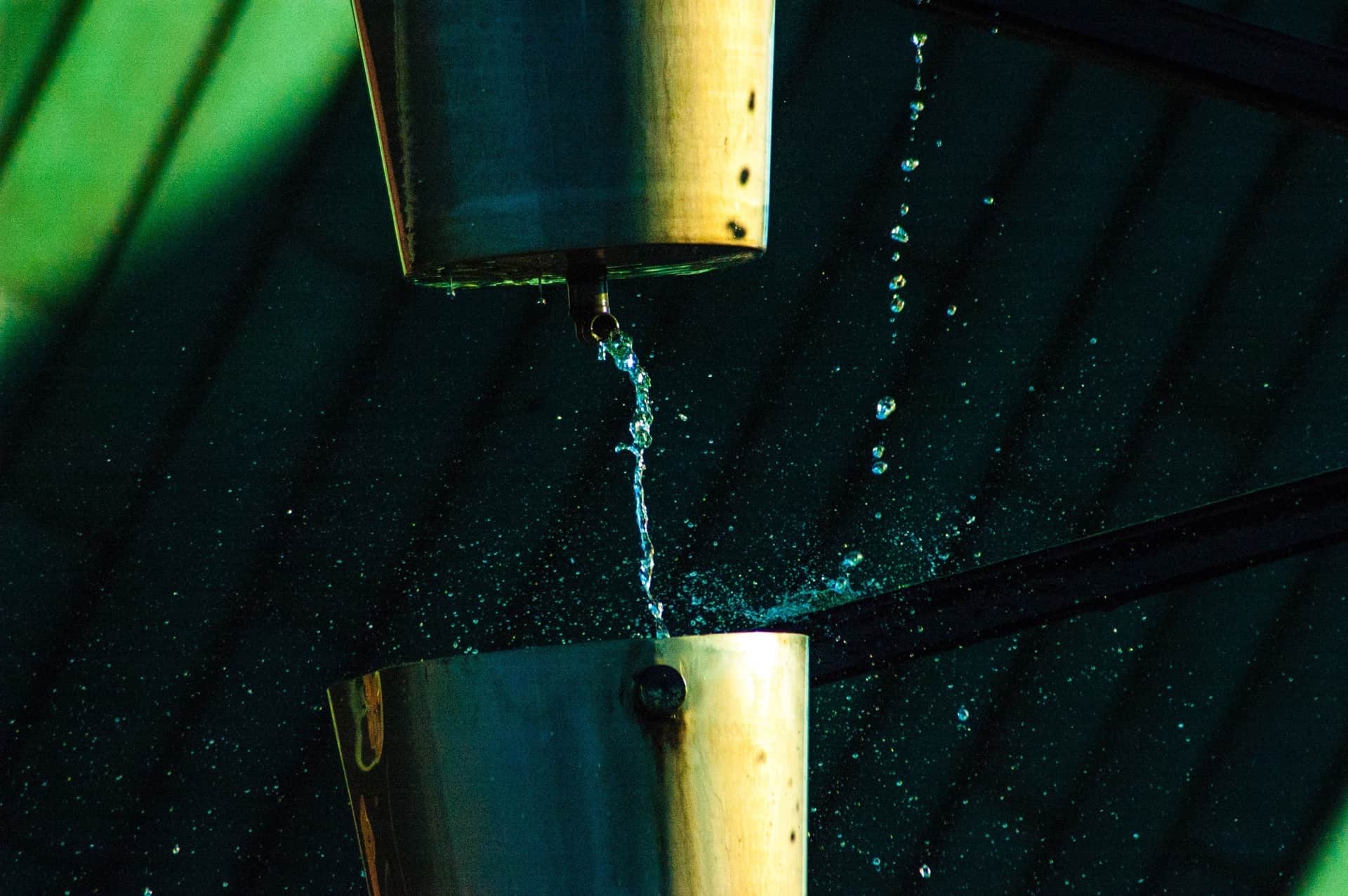 Chorro de agua cayendo de un canal metálico a otro, con cientos de gotas suspendidas sobre un fondo verde oscuro