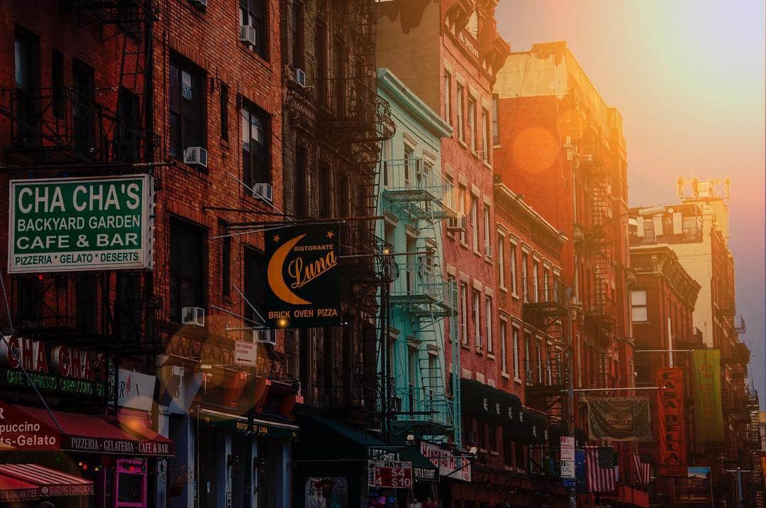 Calle de Little Italy en Nueva York, con fachadas de ladrillo, rótulos de bares y un destello de luz cruzando la imagen.