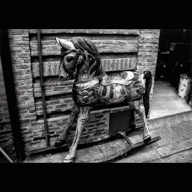 Caballito de balancín de madera apoyado contra un muro de tablones, en blanco y negro.