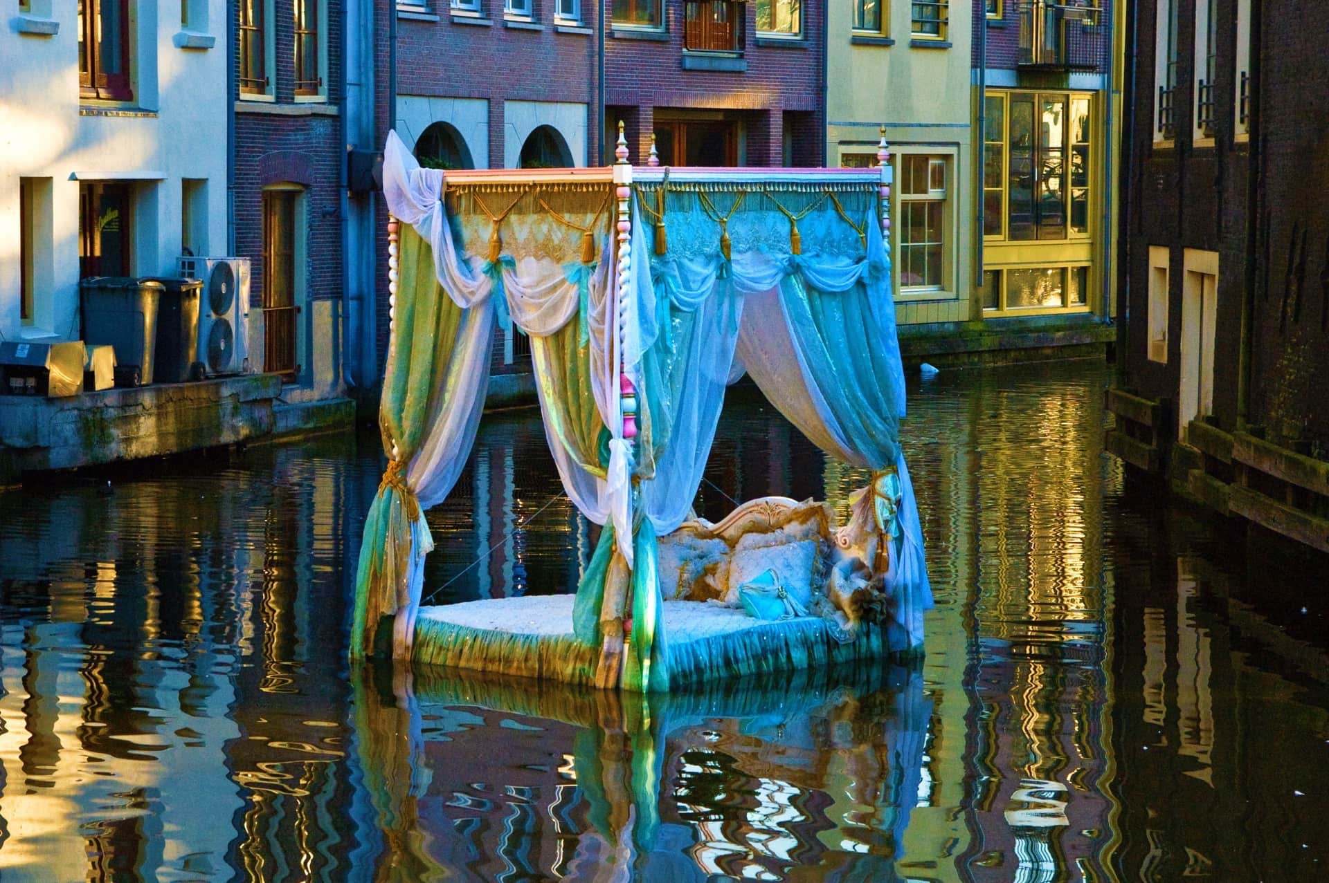 Cama con dosel y velos de gasa turquesa flotando en un canal, entre casas de ladrillo reflejadas en el agua