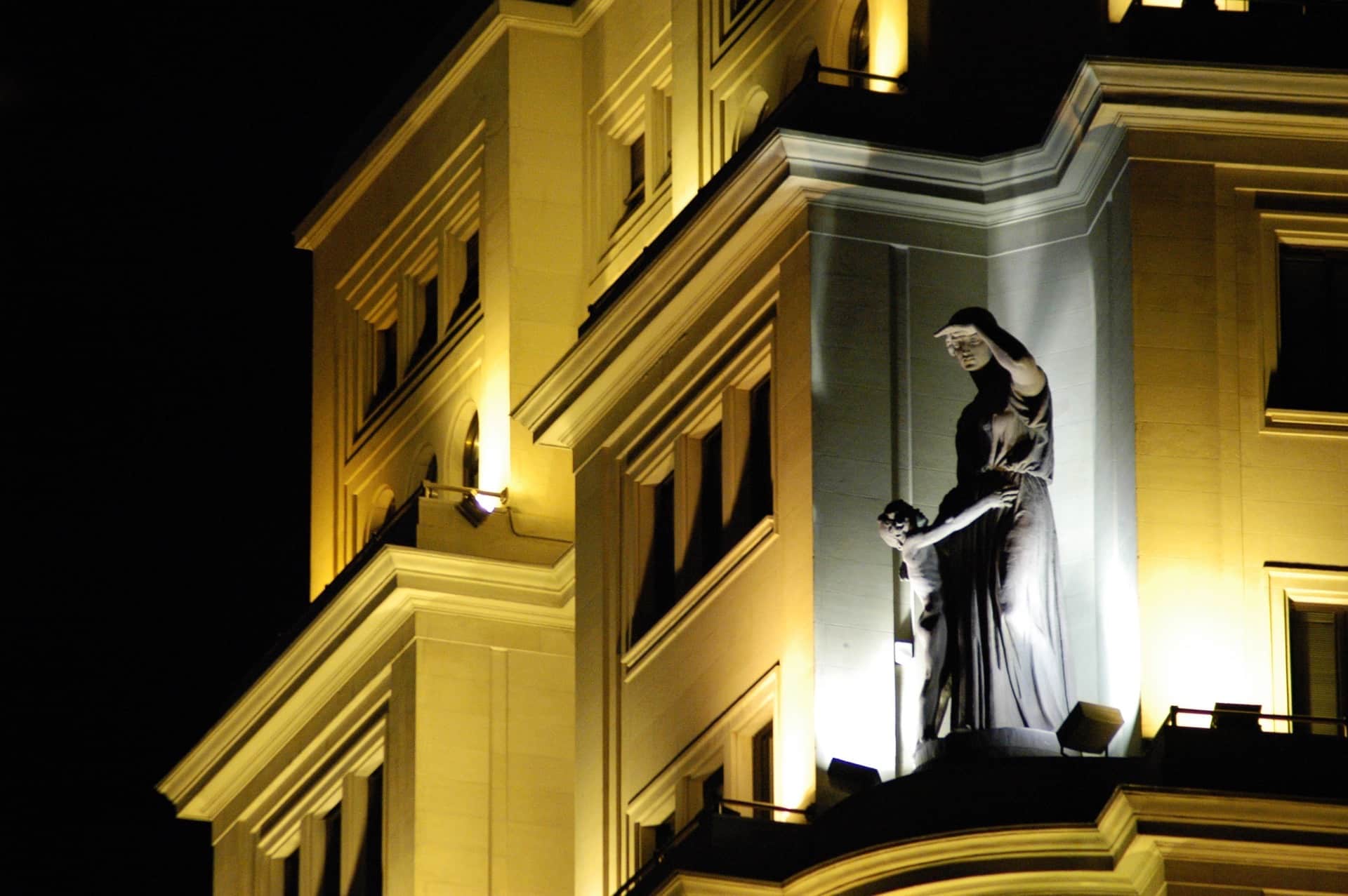 Escultura de mujer con velo, con la mano en la frente y un crucifijo, en la esquina de un edificio iluminado de noche