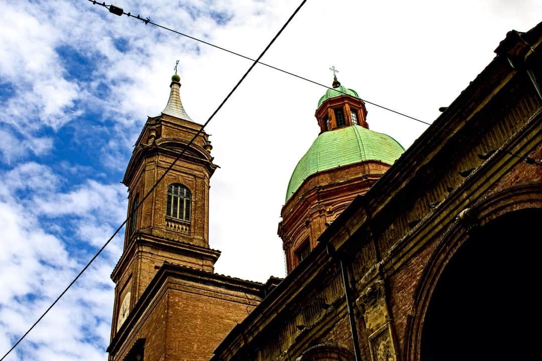Campanario y cúpula verde de una iglesia de Bolonia vistos desde abajo, con cables cruzando el cielo.