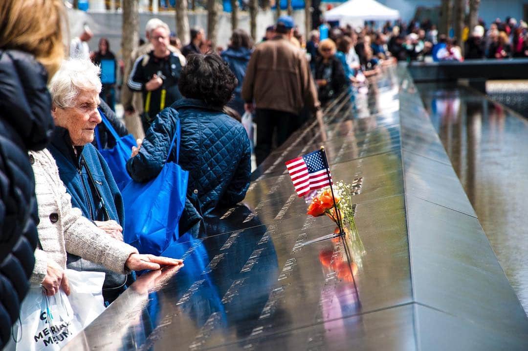 Grupo de personas asomadas a la barandilla metálica de un memorial, con una banderita de Estados Unidos apoyada en el borde.