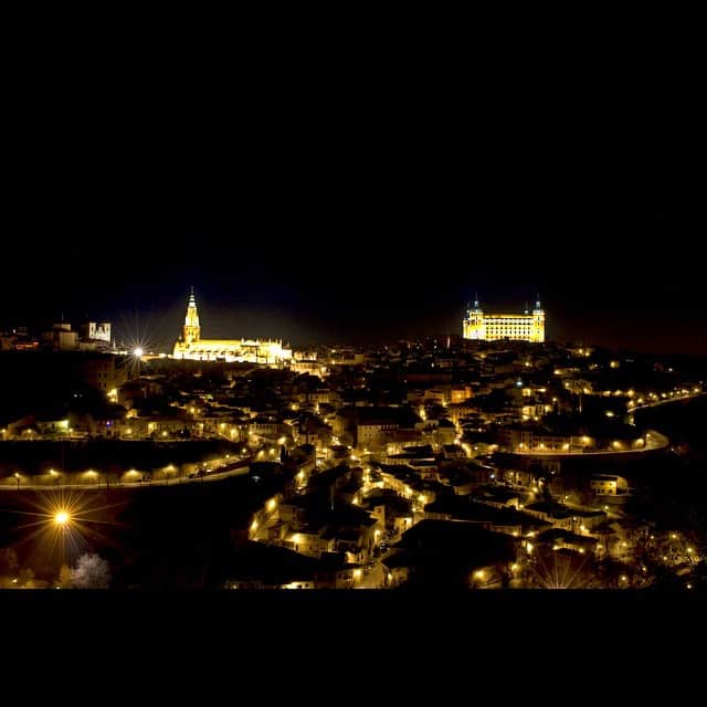 Toledo de noche, con el alcázar y el caserío iluminados en dorado sobre el fondo negro del valle.
