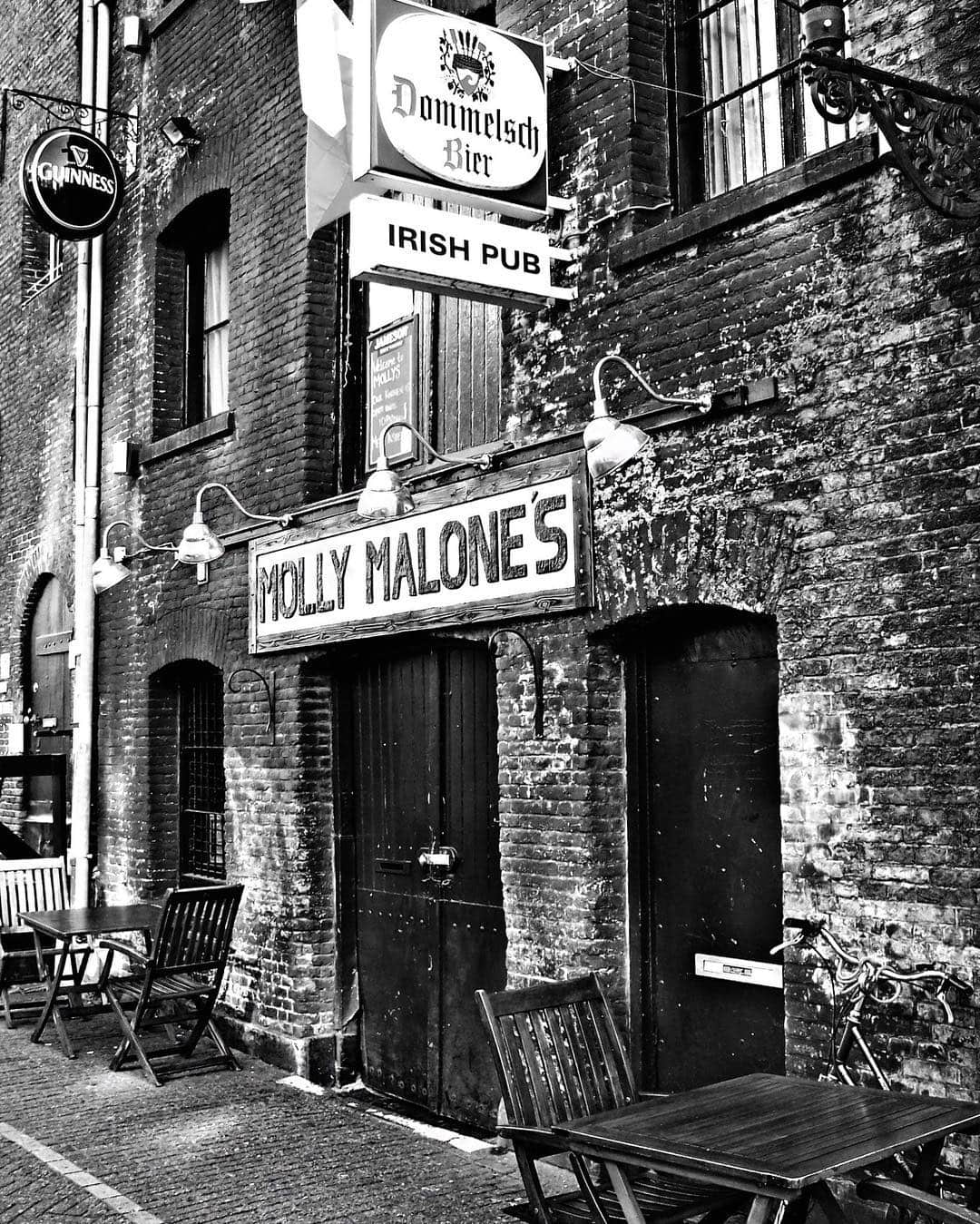 Esquina de un pub irlandés en blanco y negro, con el rótulo «Molly Malone's» y mesas vacías en la acera.