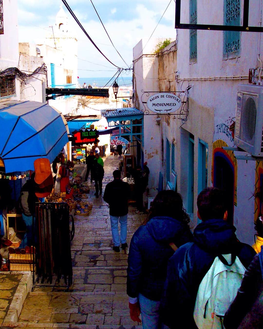Calle comercial de un zoco, con toldos y gente caminando entre casas encaladas y azules.
