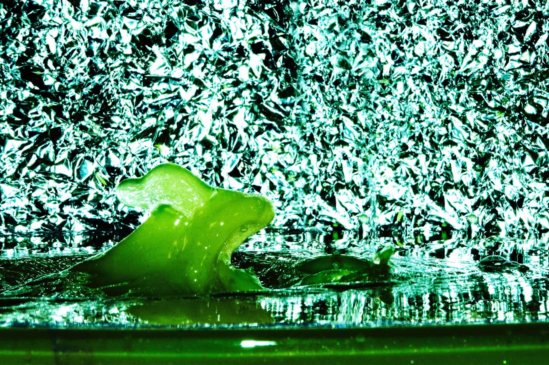 Figura gelatinosa verde con forma de hipopótamo emergiendo del agua ante un fondo de lámina metálica arrugada en verde y cian.
