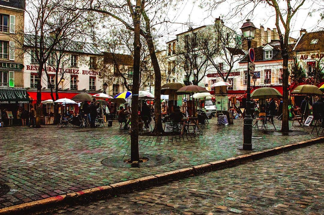 Plaza de Montmartre con terrazas, sombrillas y árboles desnudos, sobre suelo empedrado mojado.