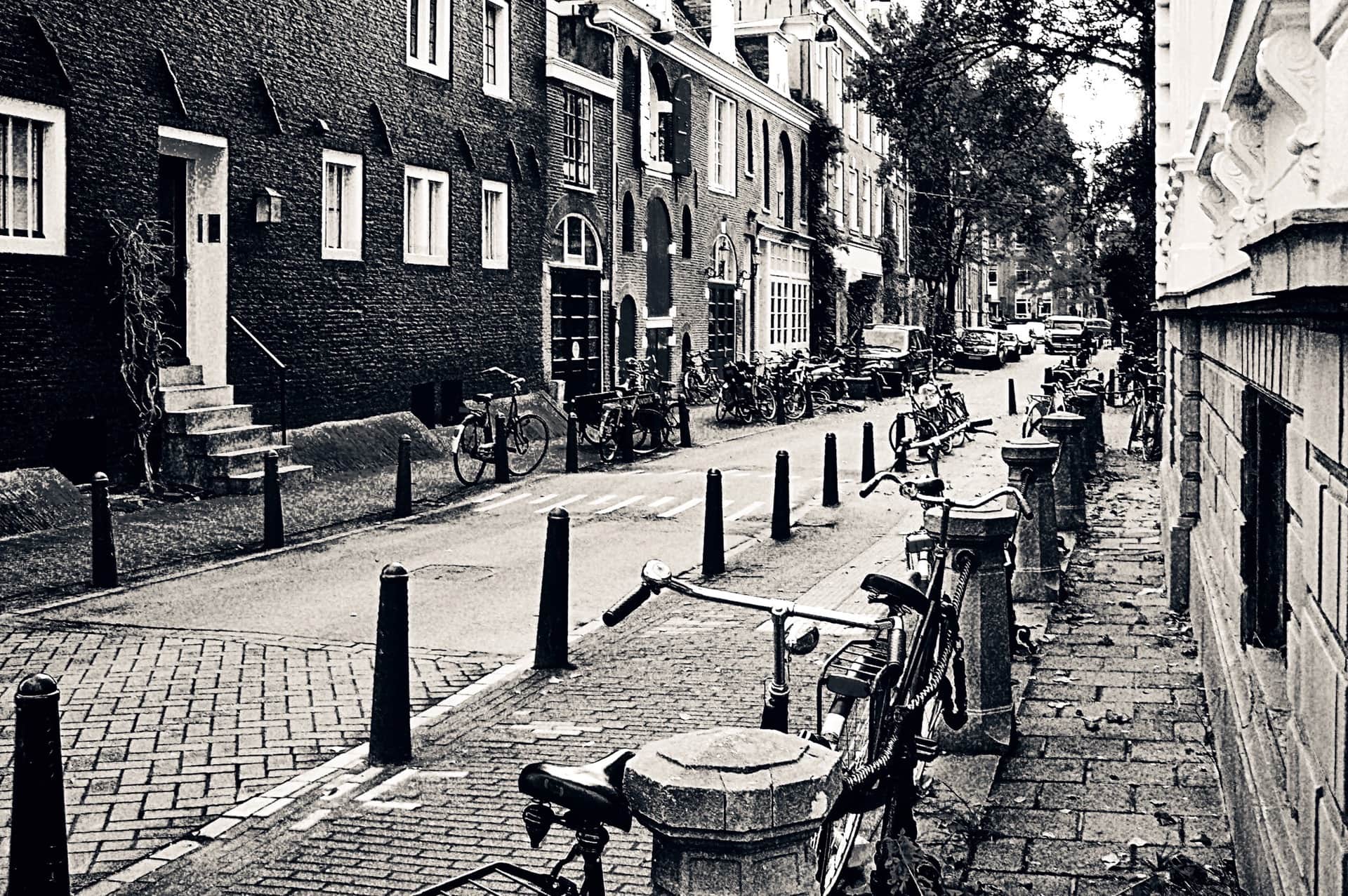 Calle estrecha con casas de ladrillo de hastiales, bicicletas aparcadas y una hilera de bolardos en la acera, en blanco y negro.