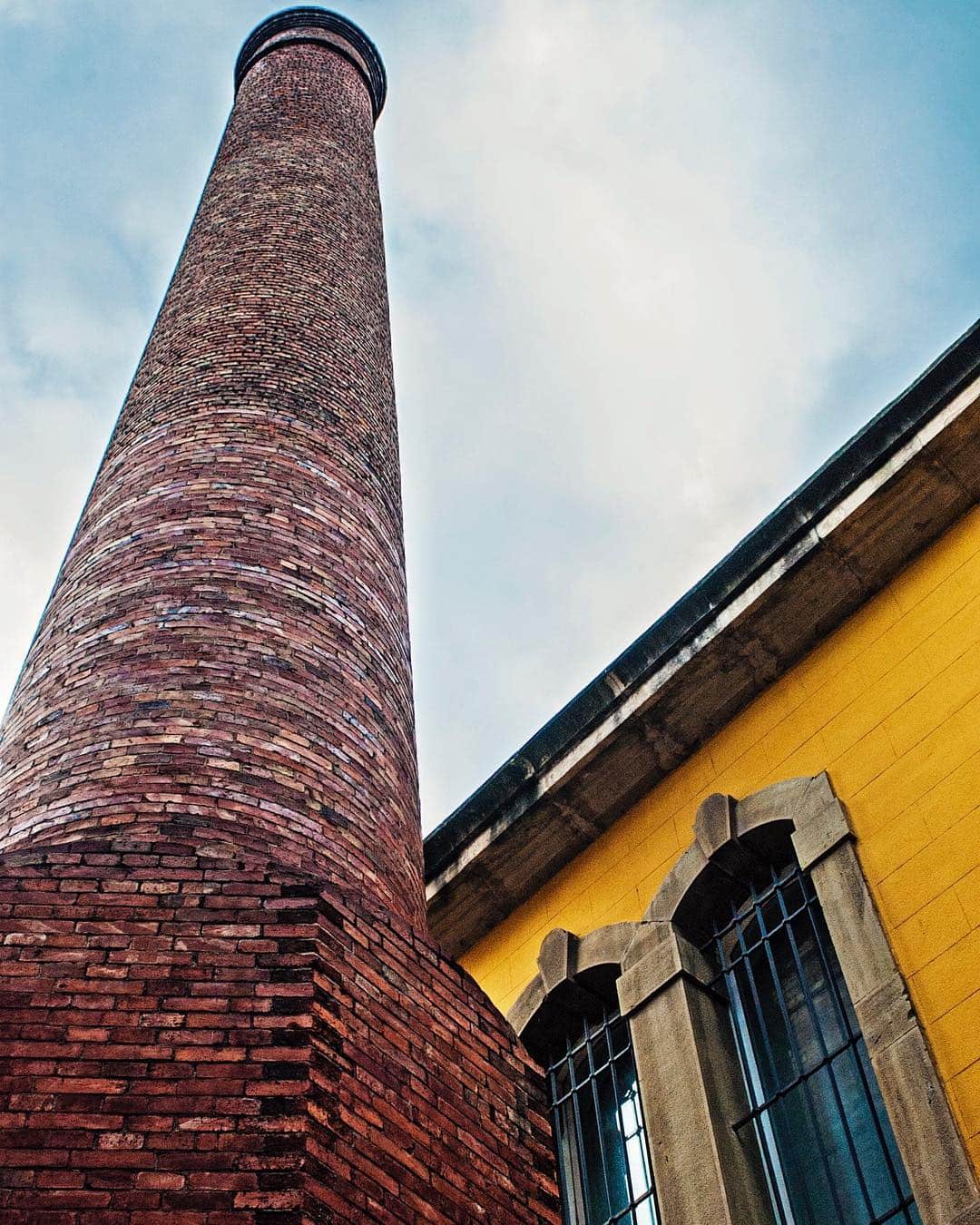 Chimenea industrial de ladrillo vista desde abajo, junto a una fachada amarilla y un cielo con nubes.