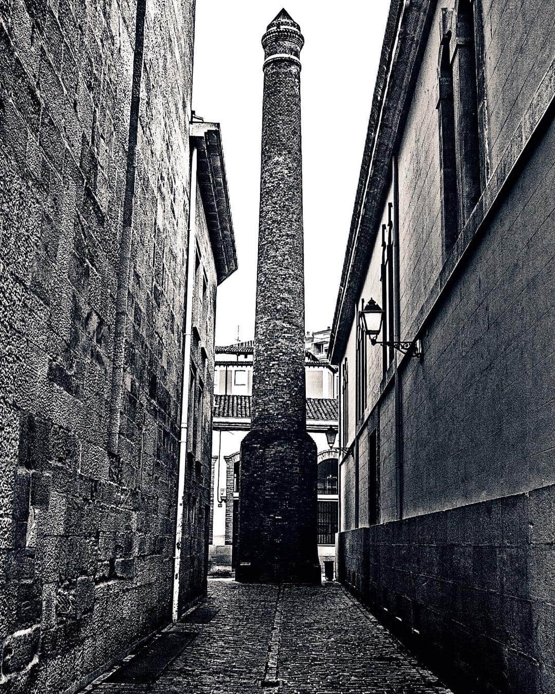 Chimenea de ladrillo al fondo de un callejón estrecho entre muros de piedra, en blanco y negro.