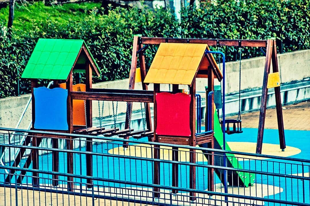 Parque infantil con estructuras de juego de colores vivos, rodeado de una valla y un seto verde.