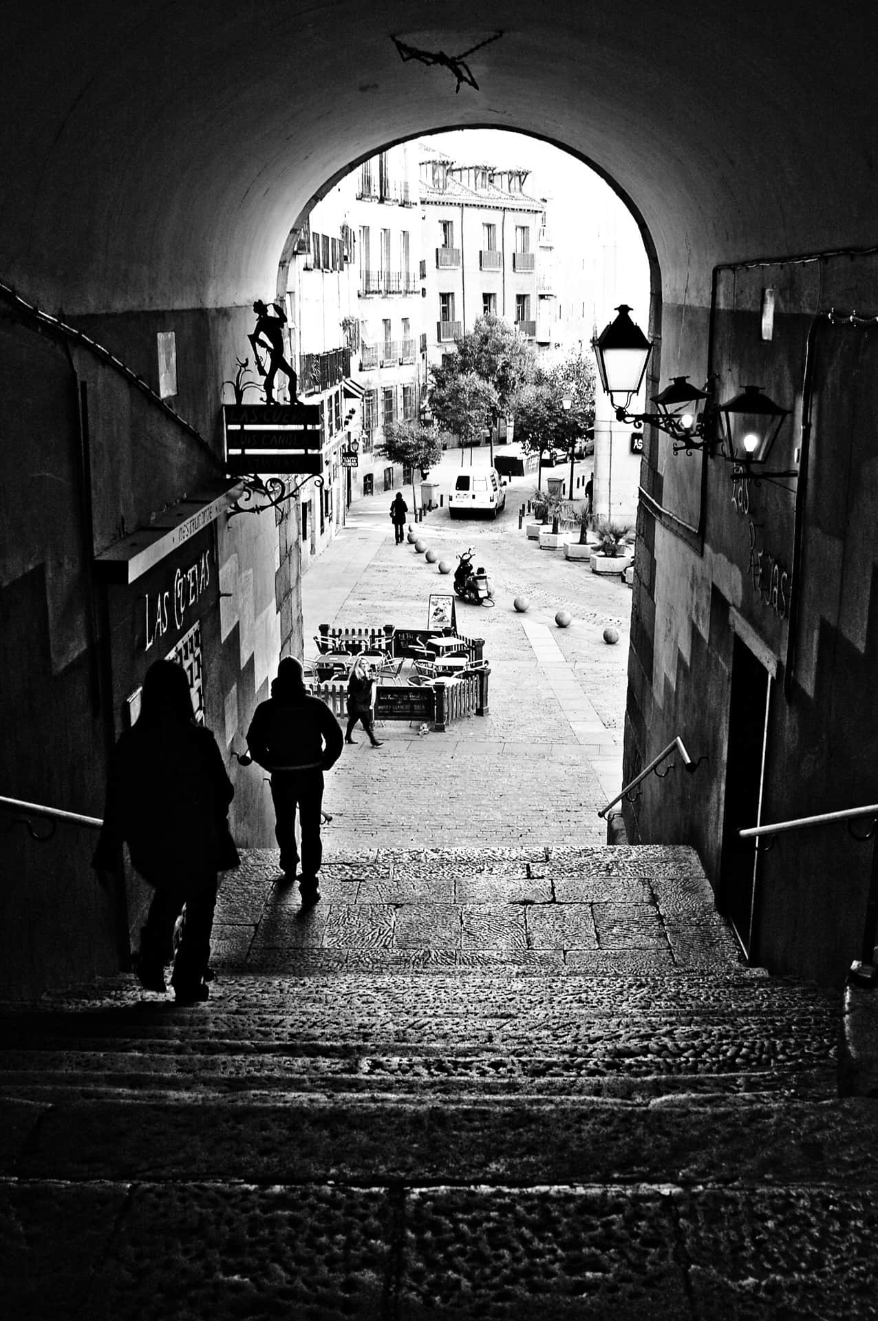Escalinata en penumbra bajo un arco de medio punto; dos siluetas descienden hacia una calle empedrada con terraza y farolas.