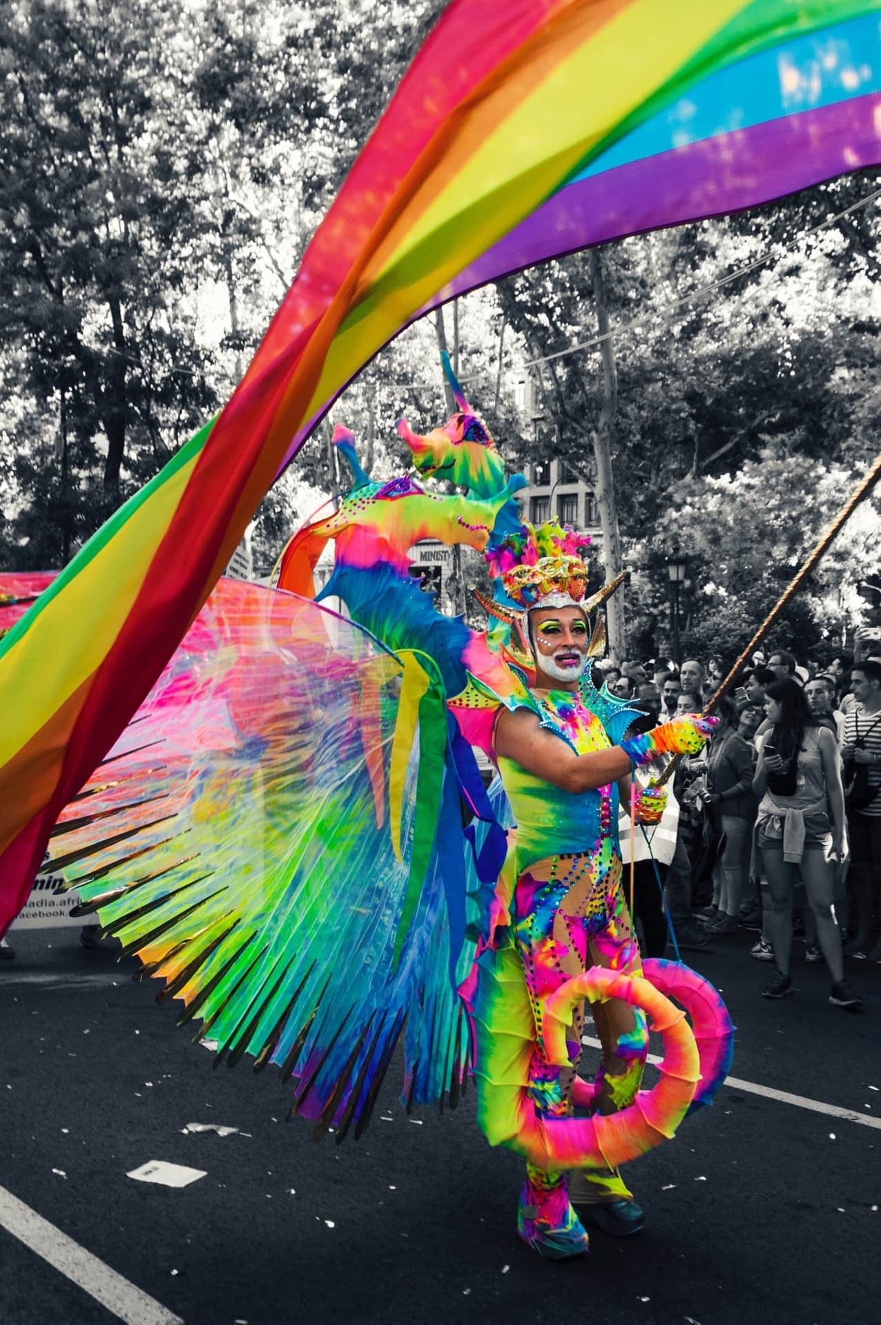 Persona con traje y tocado fluorescentes de colores sostiene una gran bandera arcoíris en un desfile; el fondo, en blanco y negro.