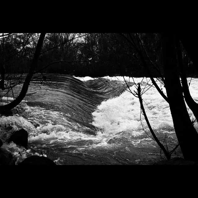 Rápidos de un río en blanco y negro, con el agua batida en blanco y ramas oscuras cruzando el encuadre.