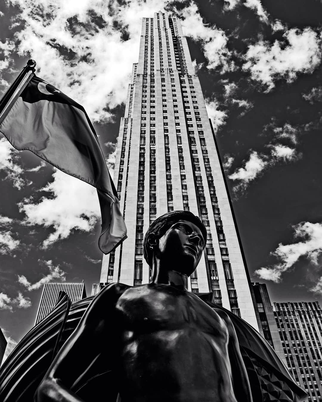 Escultura de bronce ante el Rockefeller Center, con el rascacielos elevándose al fondo, en blanco y negro.