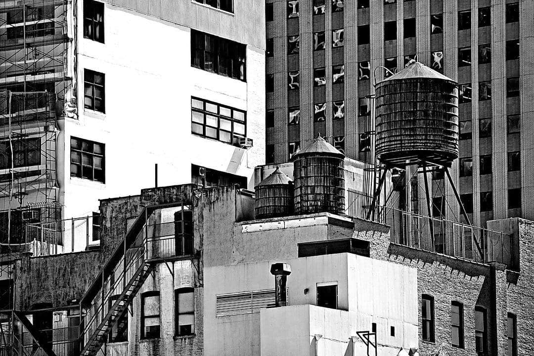 Depósitos de agua sobre las azoteas de Nueva York, entre fachadas de ventanas, en blanco y negro.
