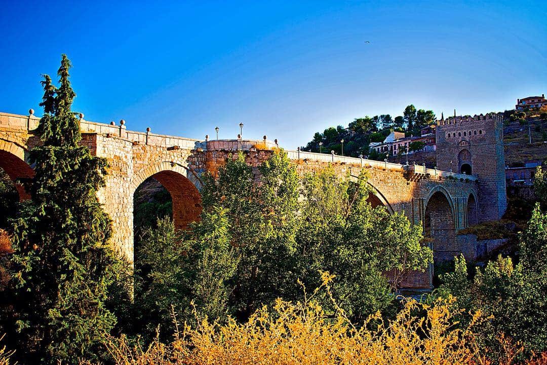 Puente medieval de piedra sobre un río, con la muralla y el caserío al fondo bajo un cielo azul.