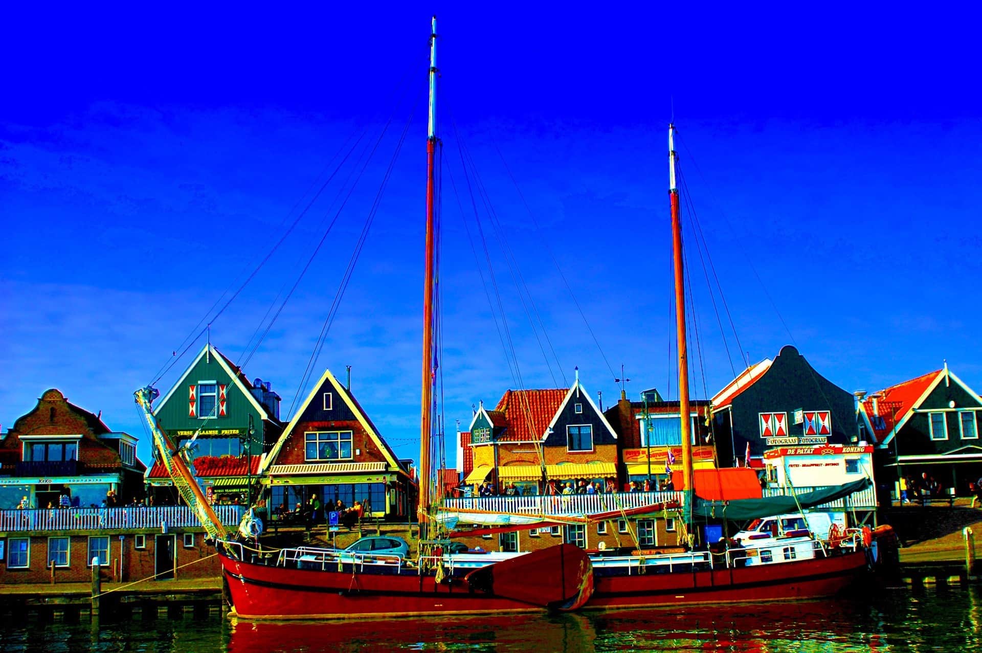 Dos veleros de casco rojo amarrados ante las casas con hastial y las terrazas de un puerto, bajo un cielo azul saturado.