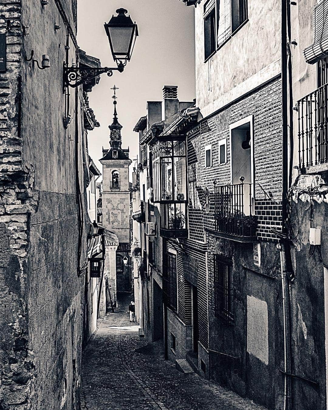 Calle estrecha en blanco y negro con una farola colgada, balcones de hierro y un campanario al fondo.