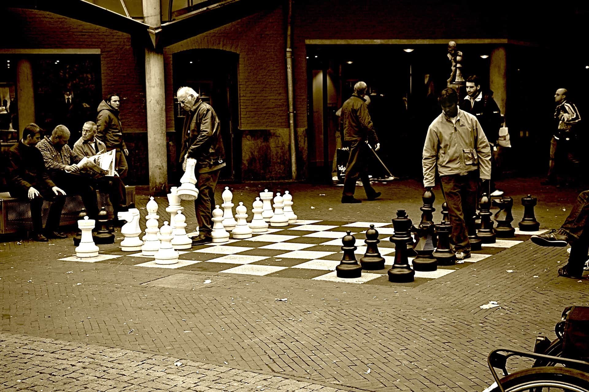 Dos hombres juegan al ajedrez con piezas gigantes en el suelo de una plaza porticada, con espectadores sentados, en sepia.