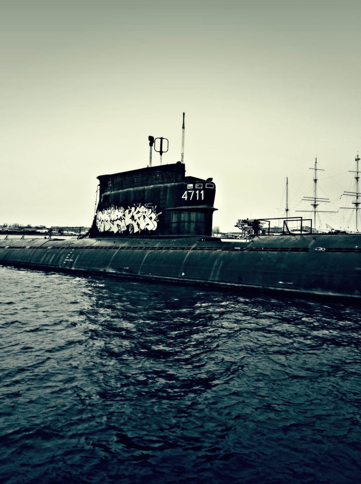 Submarino oscuro con el número 4711 y grafitis en la torre, amarrado en el agua, con mástiles de un velero al fondo.