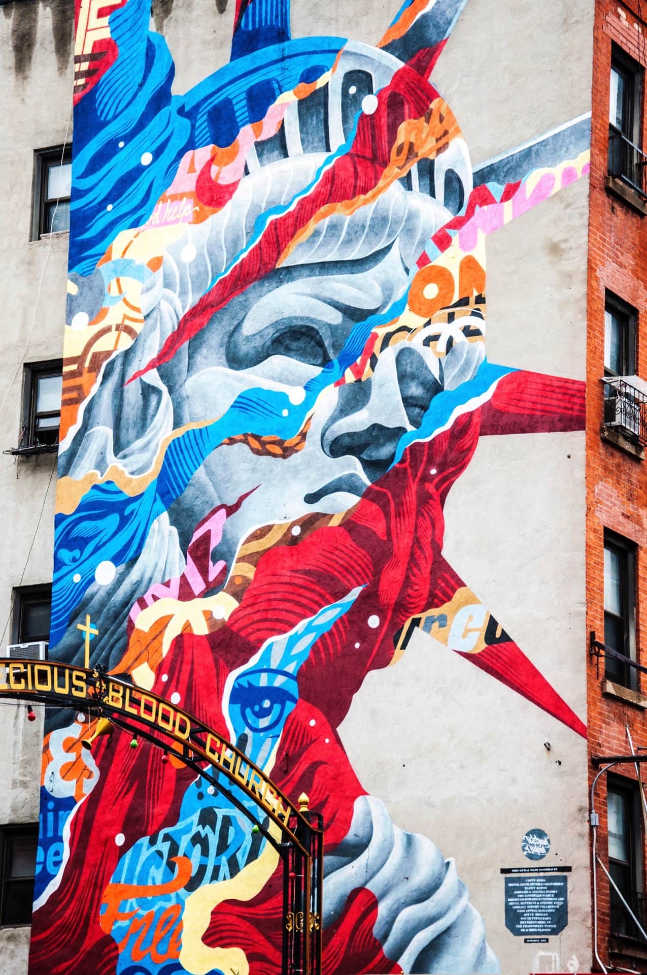 Mural del rostro de la Estatua de la Libertad, troceado en franjas rojas y azules, en la medianera de un edificio urbano.