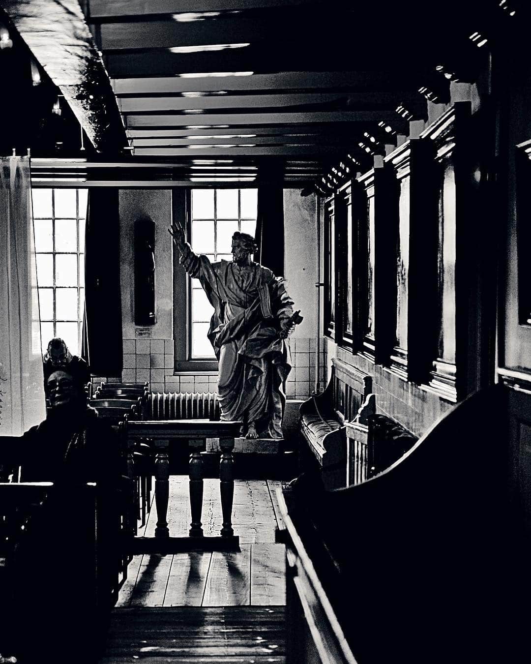 Interior de una galería en penumbra con una escultura de un apóstol junto a una ventana, en blanco y negro.