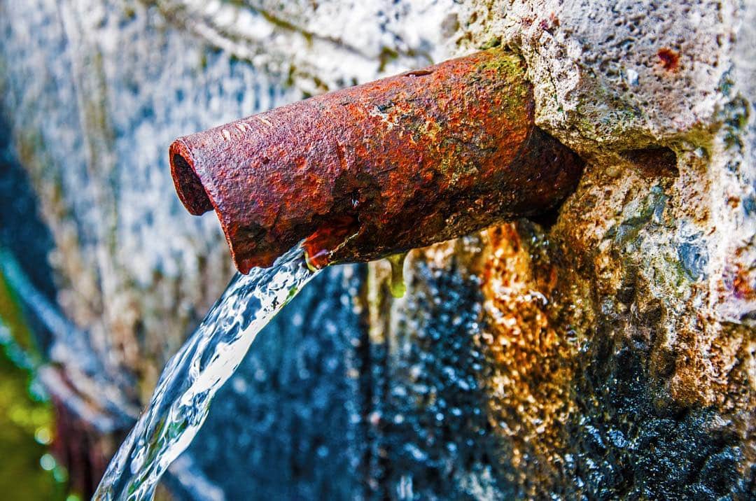Caño de hierro oxidado saliendo de un muro de piedra, con el agua cayendo en primer plano.