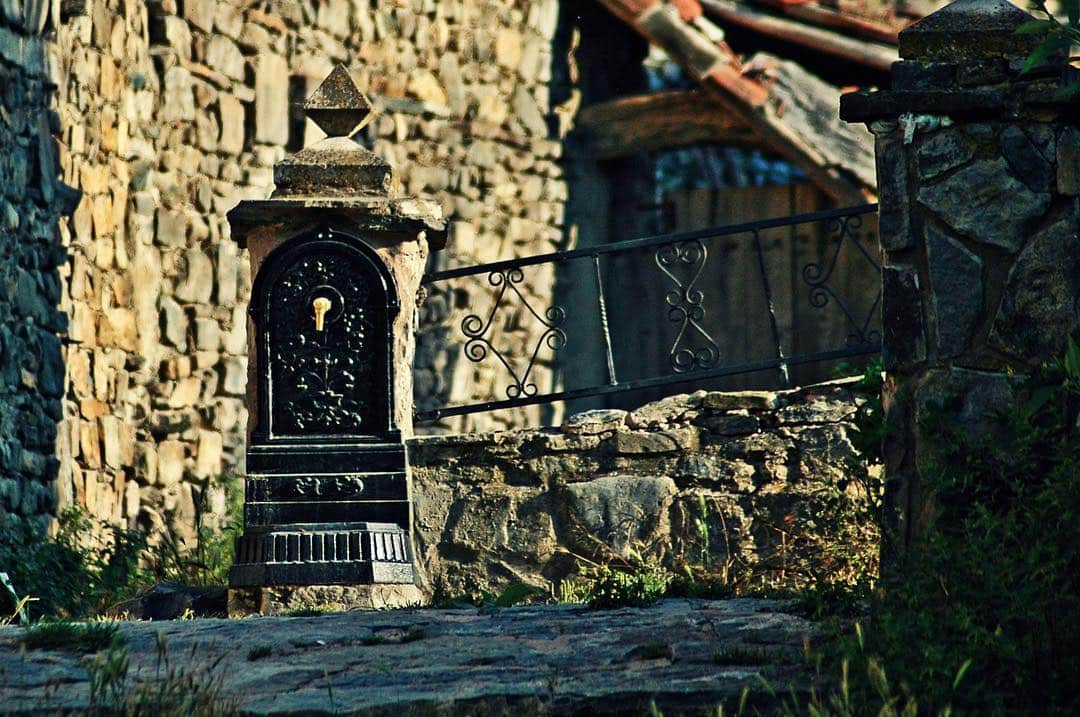 Fuente de piedra con un caño de hierro, empotrada en un muro y rodeada de vegetación.