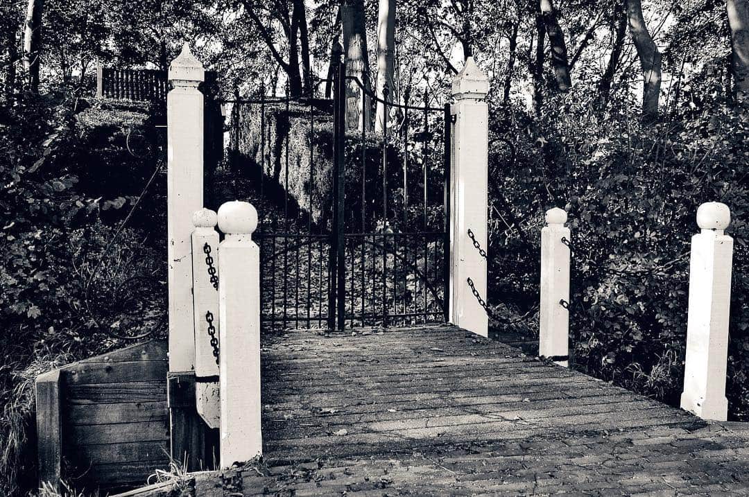 Verja de hierro abierta entre pilares de piedra, al comienzo de un puente de madera, en blanco y negro.