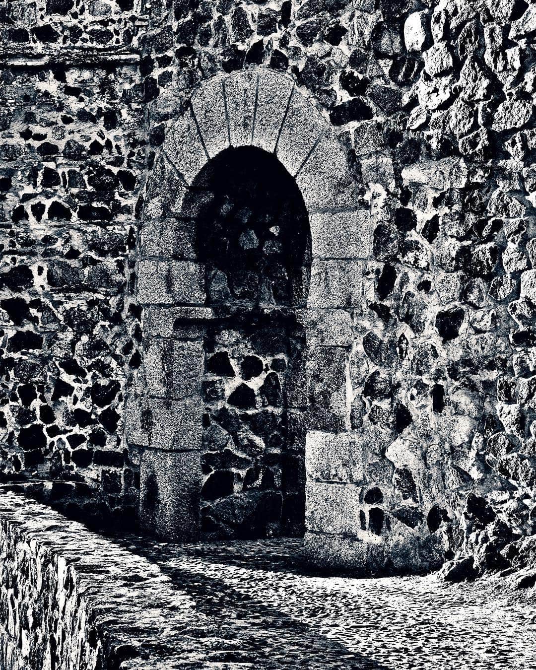Puerta en arco abierta en una muralla de piedra, con el empedrado en primer plano, en blanco y negro de alto contraste.