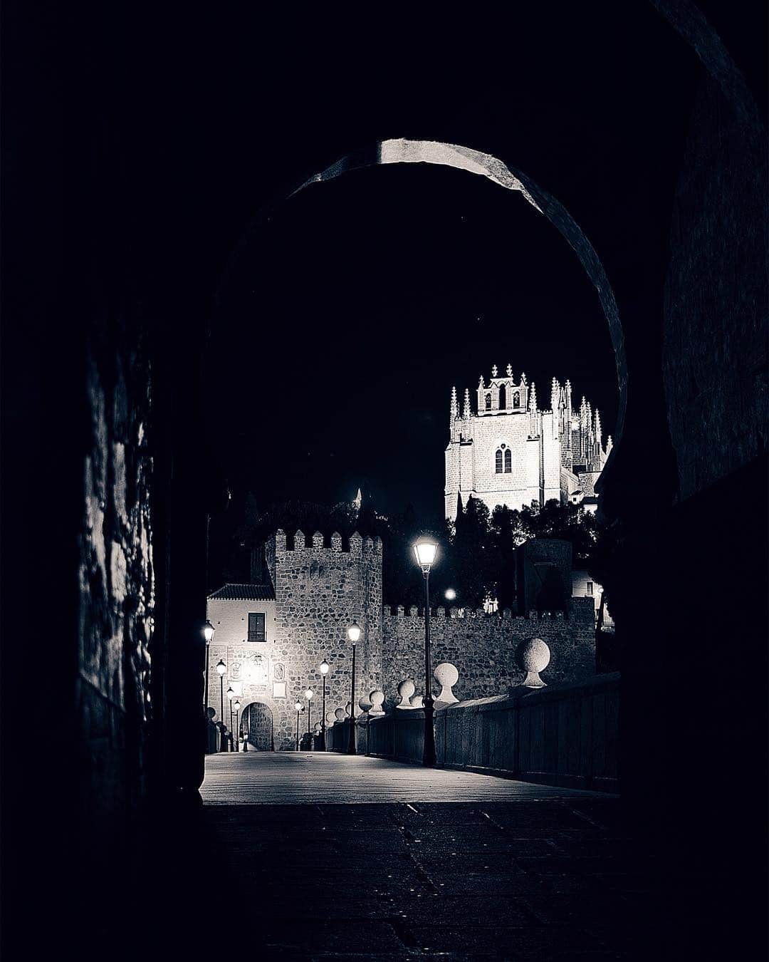 Puerta fortificada de Toledo iluminada de noche, vista a través del arco oscuro de otra puerta.