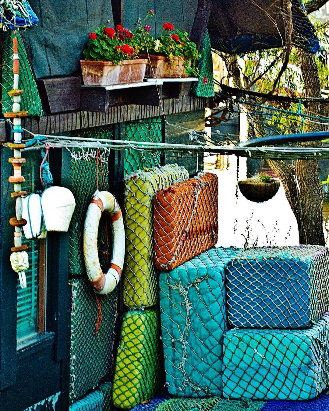 Cubierta de un barco con redes verdes y azules, un salvavidas blanco y jardineras con geranios rojos.