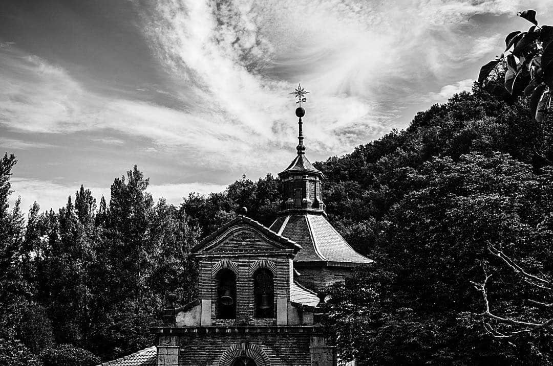 Ermita de piedra con espadaña, en blanco y negro, al pie de una ladera boscosa y bajo un cielo de nubes finas.