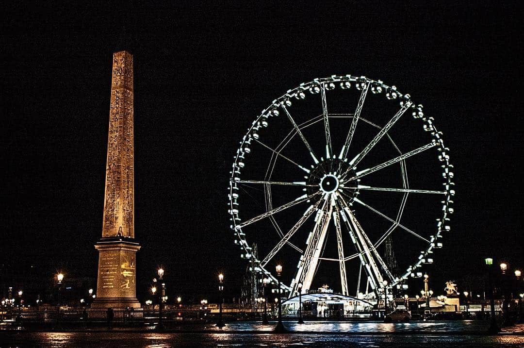 Obelisco y noria iluminada de noche en una plaza, sobre un cielo negro.