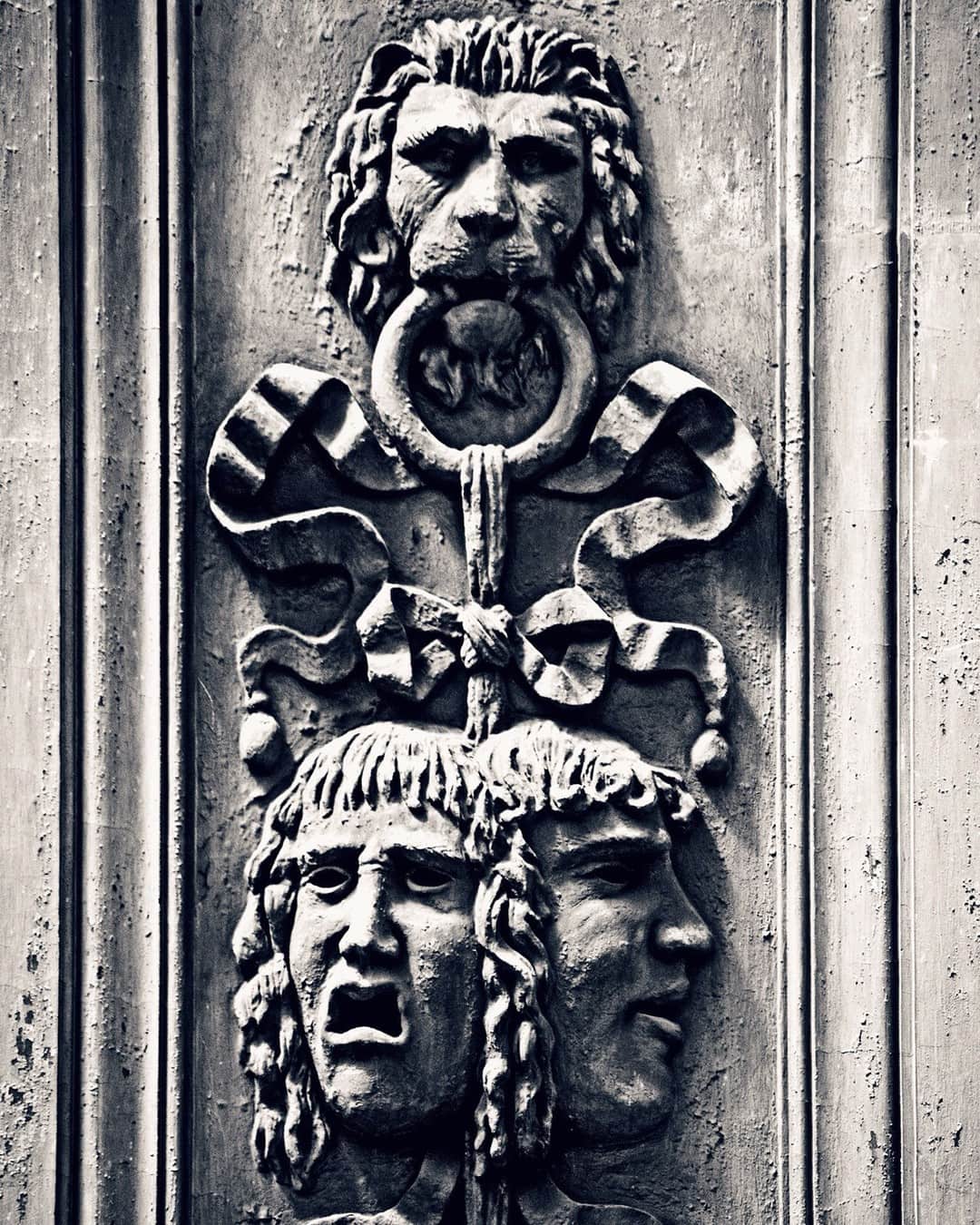 Relieve de piedra con una cabeza de león con aldaba y dos máscaras teatrales debajo, en blanco y negro.