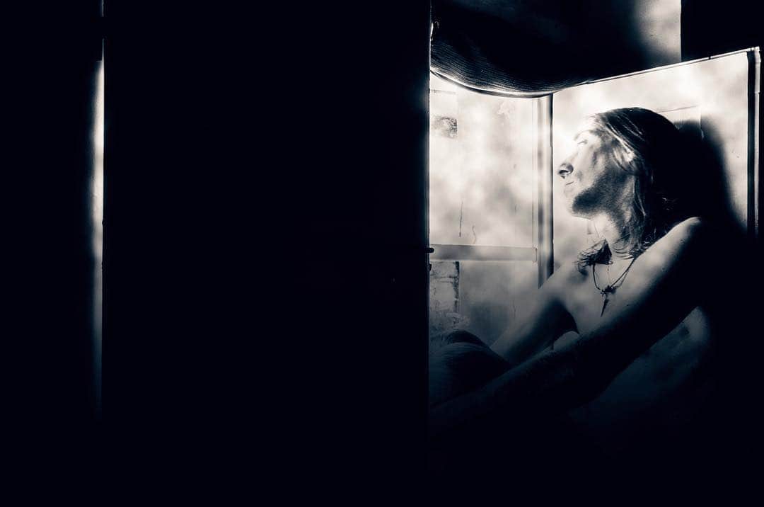 Escena en penumbra: una mujer recostada junto a una ventana, iluminada por una luz tenue, en tonos azulados casi monocromos.