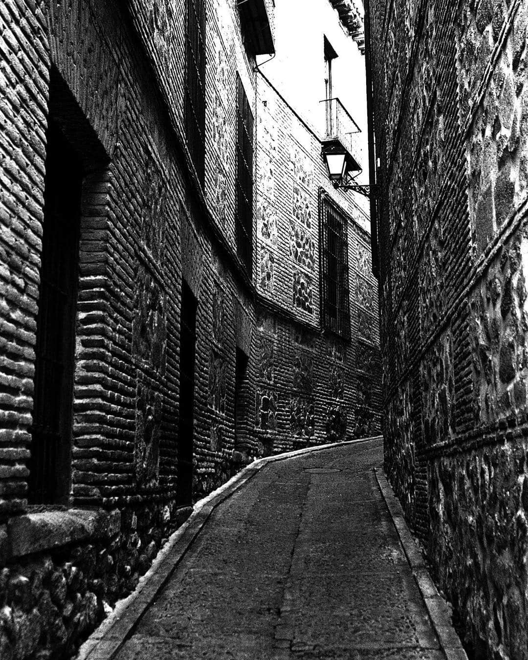 Callejón empedrado de Toledo en blanco y negro, entre muros altos de ladrillo, con una farola y un arco al fondo.