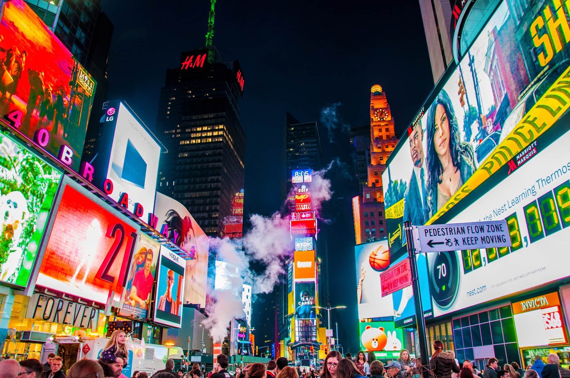 Pantallas publicitarias gigantes iluminan una multitud nocturna junto al rótulo de Broadway, con vapor saliendo del asfalto.