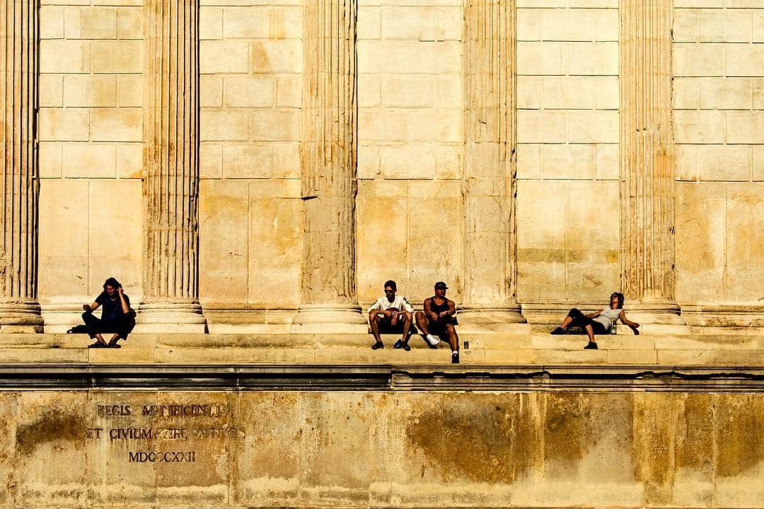 Tres grupos de personas sentadas y tumbadas al pie de un muro de piedra dorada con pilastras.