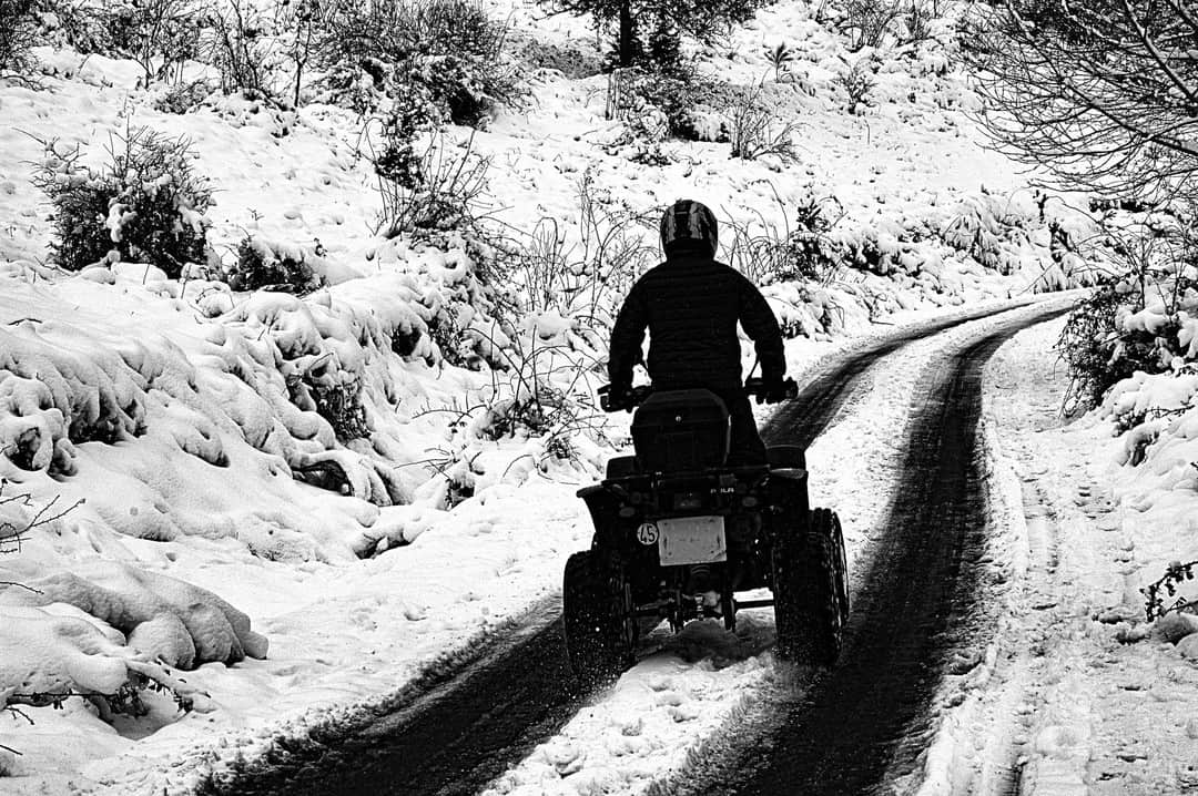Persona en quad avanzando por una carretera nevada entre matorrales, en blanco y negro.